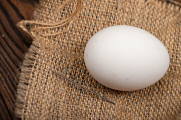 A chicken egg and a sewing needle on a homespun fabric with a rough texture.