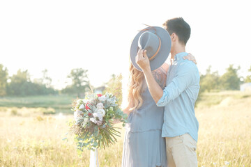 Kissing young man and woman, with a bouquet of fresh flowers in the field, in nature. A man holds a hat