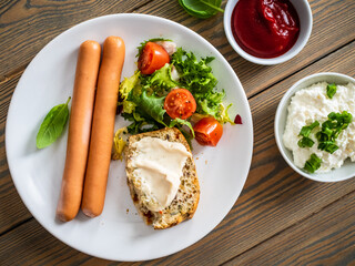 Breakfast - boiled sausages, cottage cheese, bun with mayonnaise and fresh vegetables served on wooden table
