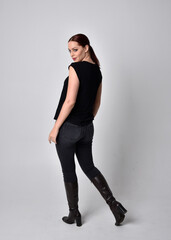 Simple full length portrait of woman with red hair in a ponytail, wearing casual black tshirt and jeans. Standing pose with back to the camera the camera, against a  studio background.