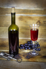 Dark green bottle and transparent wineglass with red wine and bunch of grapes on wooden plank surface, corkscrew nearby