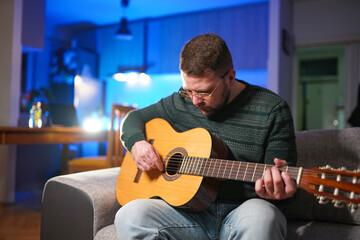 Obraz premium Young man sitting at home on sofa and playing guitar