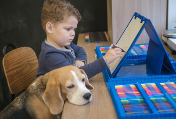 cute little boy draws with felt-tip pens sitting at the table. the beagle dog sits funny next to me