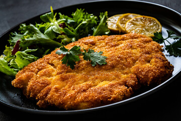 Breaded fried pork chop and fresh vegetables on black table

