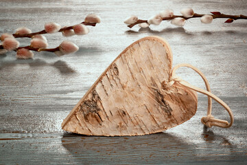 Easter decorations, toned image in desaturated earth colors. Close-up on rustic heart made from birch wood. Pussy willow twigs. Rustic wooden table with textured painted surface in counterligt.