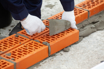 Hands in work gloves with a trowel put a red brick on the cement mortar