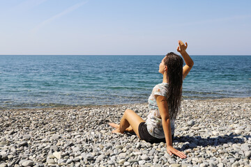 Young woman in wet clothes sits with her back alone on pebble beach and looks at horizon of the sea, on sunny day with copy space