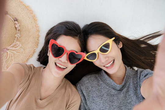 Portrait of two young Asian women wearing sunglasses in summer concept smiling and lying on the white floor to selfy take a photo. - Powered by Adobe