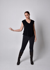 Simple full length portrait of woman with red hair in a ponytail, wearing casual black tshirt and jeans. Standing pose front on with hand reaching gestures, against a  studio background.