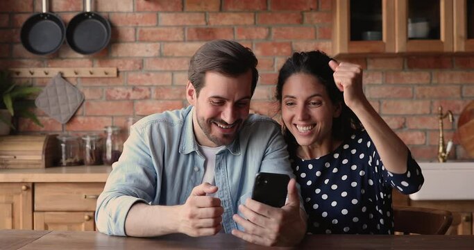 Happy couple sit at table in kitchen read received sms on smartphone device, got great commercial special offer, profit price and discount. Good news, on-line casino winners, moment of victory concept