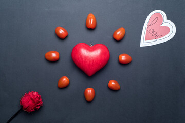 Top view on a red love heart, surrounded by small tomatoes, looking like a shining sun or a flower, with a paper heart and a red rose next to it, black background. Concept of love and valentine's day