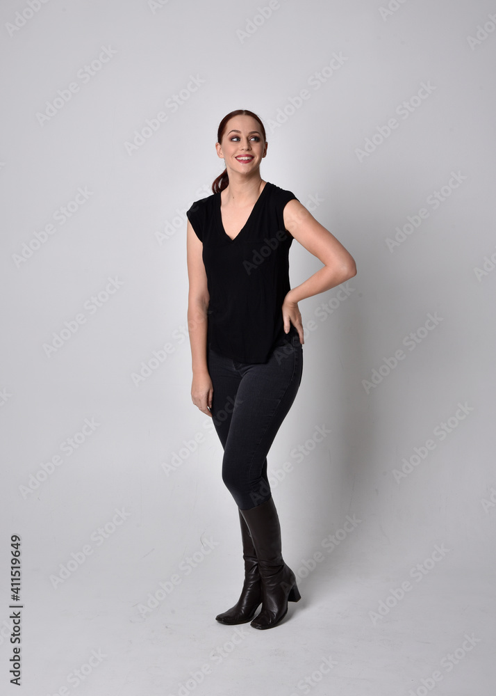 Poster Simple full length portrait of woman with red hair in a ponytail, wearing casual black tshirt and jeans. Standing pose facing front on, against a  studio background.