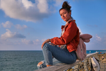 Fototapeta na wymiar beautiful latina at the sea front with red jacket and blue jeans and pink bunny suitcase