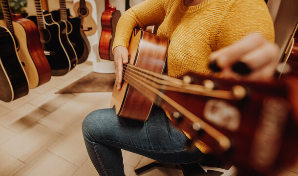 Young Woman Trying And Buying A New Wooden Guitar In Musical Instrumental Shop Or Store