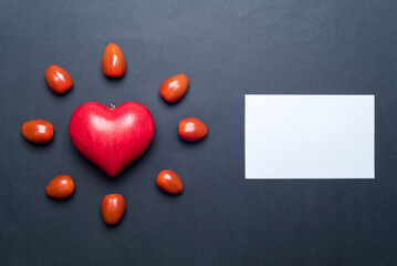 Red love heart, surrounded by small tomatoes, looking like a shining sun or a flower, with a blank gift card next to it, black background. Concept of love and valentine's day