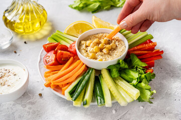 Female hand dipping carrot stick into hummus. Big plate with colorful and vitaminized vegetarian food