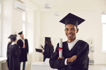 Portrait of a smiling African American graduate standing with a diploma and with folded arms and looking at the camera. Guy poses against the background of his classmates. Banner. Place for text. - obrazy, fototapety, plakaty