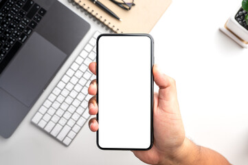 Top view of people hand holding smartphone with white blank screen over the  white table with smartphone, laptop,keyboard,notebook,pen,small tree and glasses.Flat lay design.