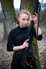blonde woman in black suit and corset with shotgun stays in front of tree trunk in the autumn forest