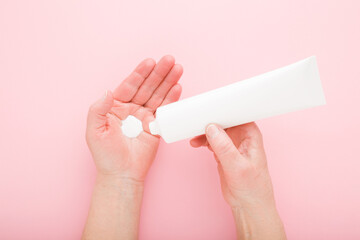 Mature woman hands using anti aging moisturizing cream on light pink table background. Pastel color. Holding white tube. Care about soft body skin. Daily beauty product. Closeup. Top down view.