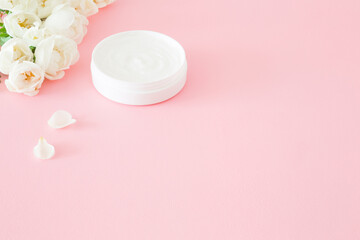 Opened white jar of natural herbal cream for women on light pink table. Pastel color. Beautiful roses. Fresh flowers. Care about clean and soft face, hands, legs and body skin. Empty place for text.