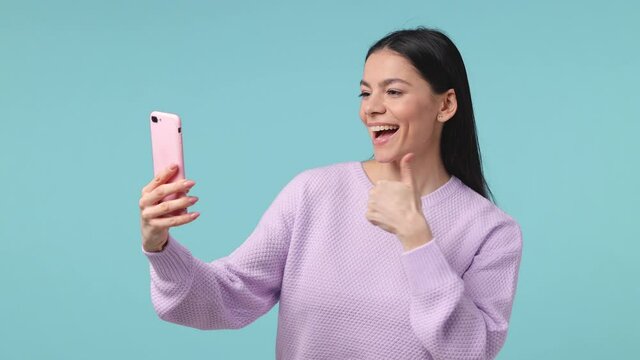 Funny young latin woman in violet sweater isolated on blue background studio. People lifestyle concept. Doing selfie shot on mobile phone showing victory sign thumb up put hand on cheek send air kiss