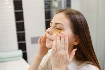 Caucasian woman applying hydrogel patches on under eye area, looking at mirror. Happy young lady...