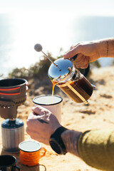 Hipster millennial man with hand tattoos prepare specialty coffee in french press, use camping stove and enamel cup. Good filter coffee during camping trip