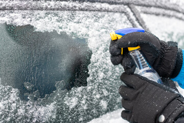 Vereiste Autoscheibe wird mit einem Enteiser-Spray eingesptüht. Wintereinbruch mit Eisregen. Hände in Winterhandschuhen und Enteiser Sprühflasche.