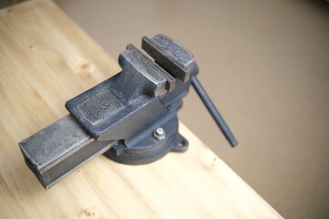 Top view a black vise with a screw grip and a cast-iron frame is mounted on a wooden countertop, in a machine shop. The wooden background is blurred.