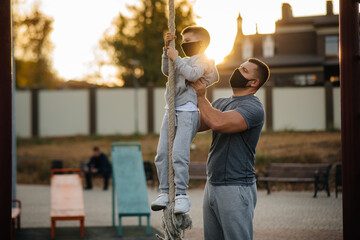 Fototapeta na wymiar A father helps his son climb a rope on a sports field in masks during sunset. Healthy parenting and healthy lifestyle