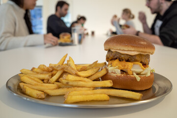 Selective focused at cheese burger beef and fried potatoes, French fried on metal tray and white cafeteria table, and blur background of people eating and having lunch together in white canteen.  