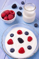 breakfast. On the lavender blue wooden table, in the foreground the bowl with the white yogurt cream with raspberries, blueberries and blackberries, in the background the yogurt jar and fresh berries.