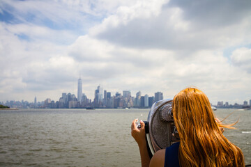 A young woman with red hair is looking at the cityscape of New York City through a binocular on Liberty Island