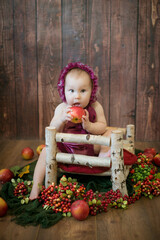 Little sweet girl up to one year old in a beautiful burgundy velvet suit with red juicy apples on a crib made of wood. Healthy lifestyle 
