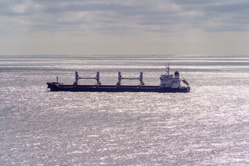 Obraz premium Large industrial ship sailing in the distance with silver waves from the sunlight and horizon with clouds in the background.