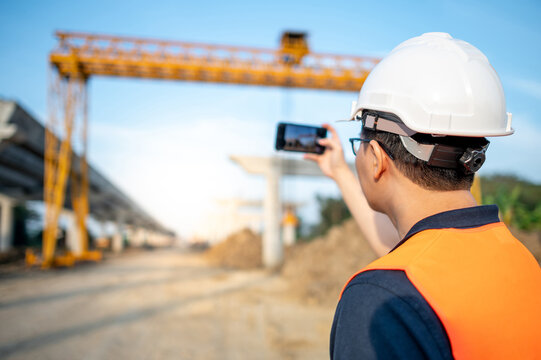 Smart Asian worker man or male civil engineer with protective safety helmet and reflective vest using using smartphone for taking photo at construction site.