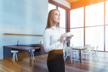 Attractive business woman drinking coffee at corporate kitchen in skyscraper office. Modern kitchen set and facilities for employees. Caucasian elegant model wearing in formal wears.