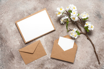 Blank wedding card mockup in brown envelopes and cherry blossom branch (artificial). Beige grunge background. Spring stationery still life. Top view, flat lay.