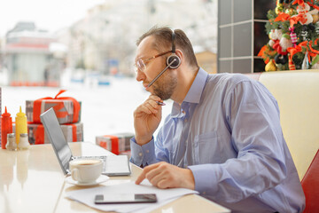 A middle-aged caucasian man with a headset in eyewear focused at the laptop screen with a pen in his hand. He works during a coffee break with a laptop, mobile phone and papers on the table sitting in