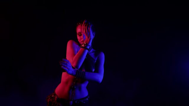 An attractive woman dances an oriental dance on a black smoky background in the studio, makes beautiful movements with her hands, she is illuminated by light.