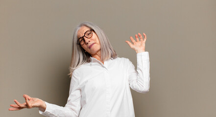 senior woman shrugging with a dumb, crazy, confused, puzzled expression, feeling annoyed and...