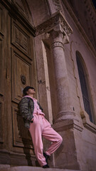 Fototapeta na wymiar latina woman with black hat leaning against a large wooden door of an old building, wearing pink sport pants and camouflage jacket