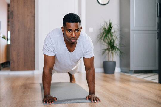 Front view of focused African-American man doing push-up on floor at bright domestic room, looking away. Concept of sport workout training at home gym.