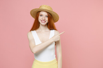 Young smiling nice caucasian redhead woman ginger long hair in straw hat summer clothes pointing fingers aside on copy space workspace area mock up isolated on pastel pink background studio portrait