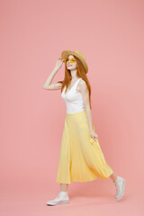 Full length body young stylish cute smiling redhead ginger woman in straw hat glasses summer clothes yellow pleated skirt walking going look aside isolated on pastel pink background studio portrait.