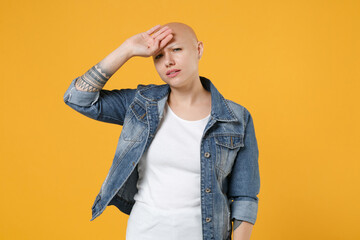 Young bald exhausted tired sick ill woman 20s without hair in casual denim jacket white t-shirt looking camera put hand on forehead head have headache isolated on yellow background studio portrait.