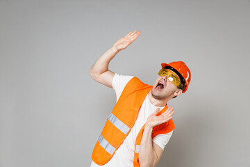 Young employee scared man in orange vest protective helmet hardhat cover with hands show palms look up isolated on grey background studio. Instruments for renovation apartment . Repair home concept.