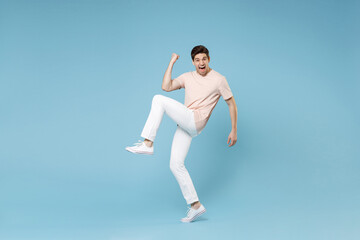 Fototapeta na wymiar Full length of young caucasian happy excited successful student man in beige t-shirt white pants do winner gesture clench fists, raise up leg look camera isolated on blue background studio portrait.