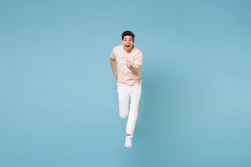 Full length of young excited energetic student man 20s in beige t-shirt white pants do winner gesture clenching fists in air jumping high running isolated on blue color background studio portrait.